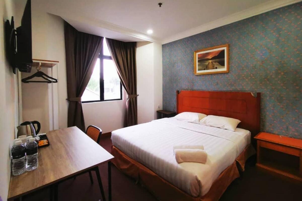 A hotel room with a bed, a desk with a chair, a table with bottles, a TV mounted on the wall, and a painting on the wall.