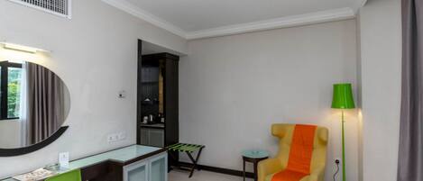 Habitación de hotel con un escritorio, una silla verde, un sillón amarillo con una manta naranja, una mesita y una lámpara de pie verde, además de un espejo.