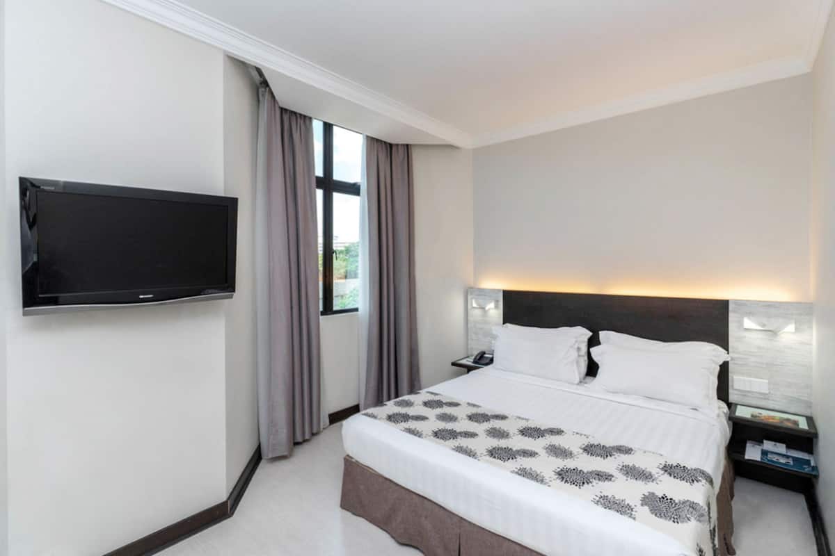 A hotel room with a bed, a flat-screen TV mounted on the wall, a nightstand with a lamp, and a window with curtains.