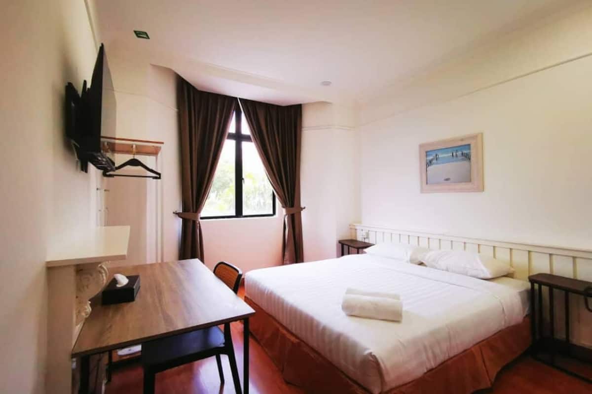 A hotel room with a bed, a desk, a chair, a TV mounted on the wall, and a window with curtains.