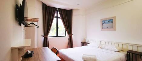 A hotel room with a bed, a desk, a chair, a TV mounted on the wall, and a window with curtains.