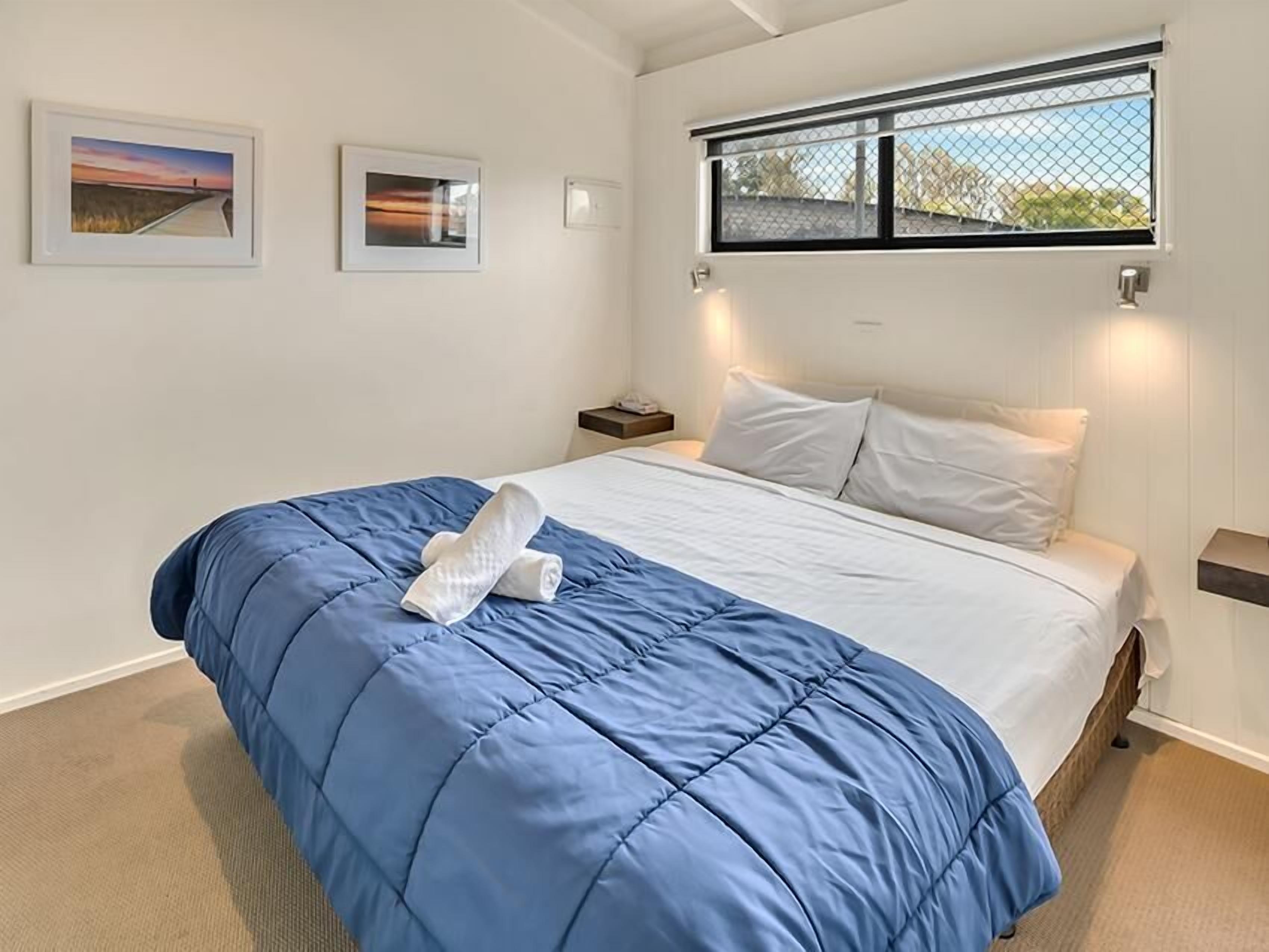 A neatly made bed with a blue comforter and white pillows, a nightstand with a lamp, and framed pictures on the wall.