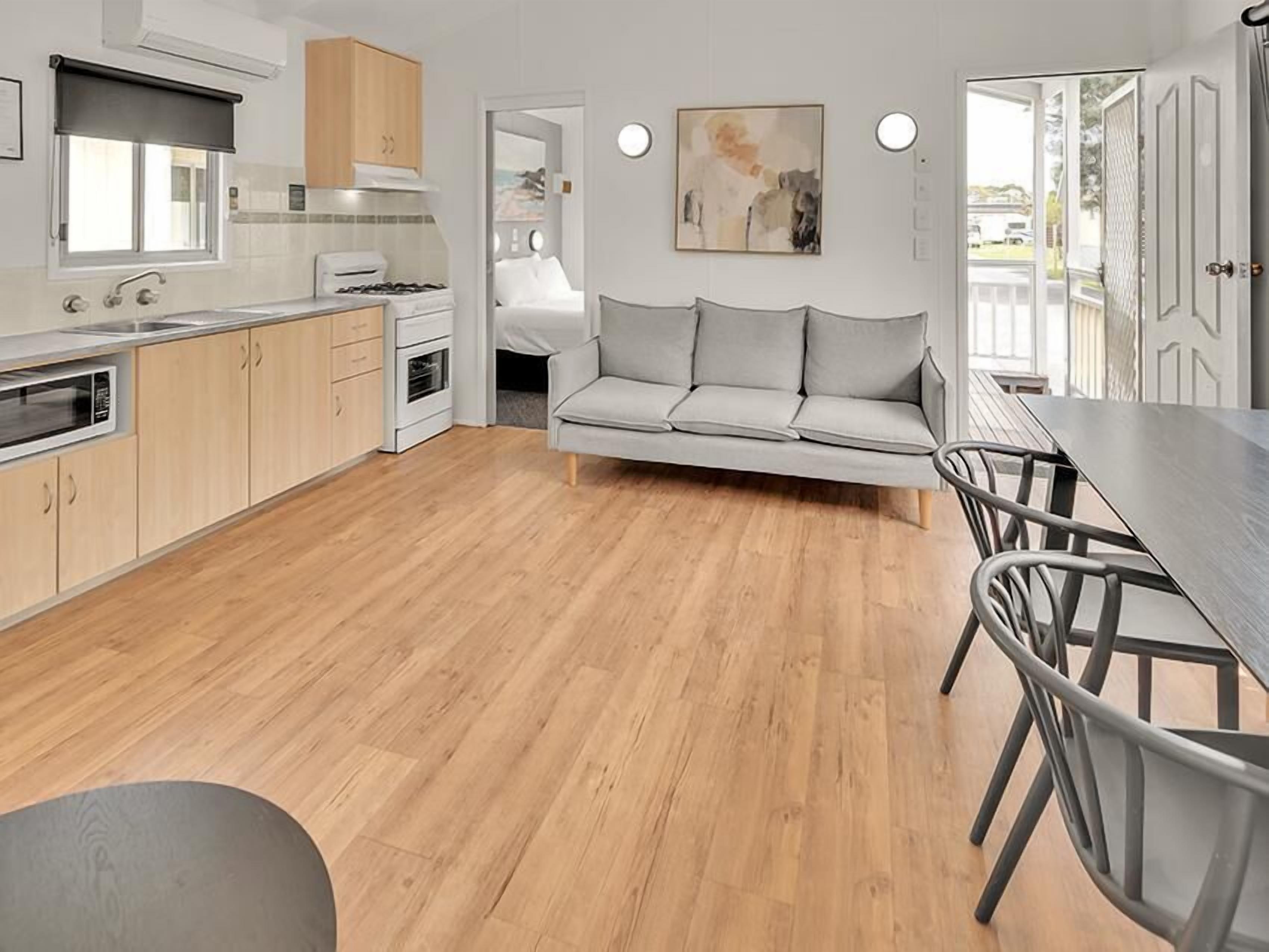 Une cuisine moderne avec un plancher en bois, un coin repas avec un canapé gris et des chaises en métal, et une vue à travers une porte ouverte.