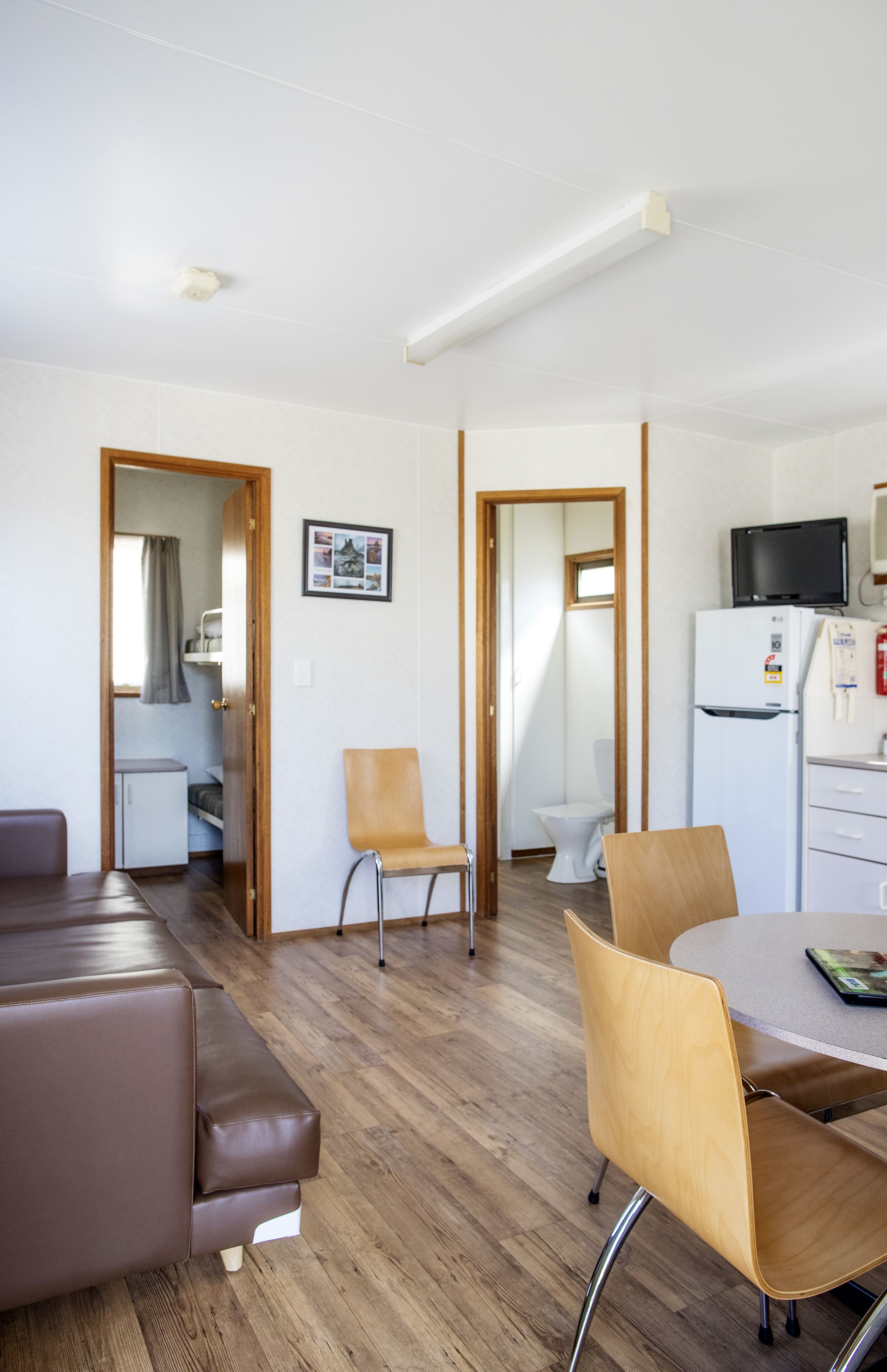 A modern, well-lit living room with a brown leather sofa, wooden chairs, a dining table, and a kitchen area with a refrigerator and microwave.