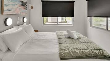 A hotel room with a bed, two windows with blinds, a painting on the wall, and two wall-mounted lights.