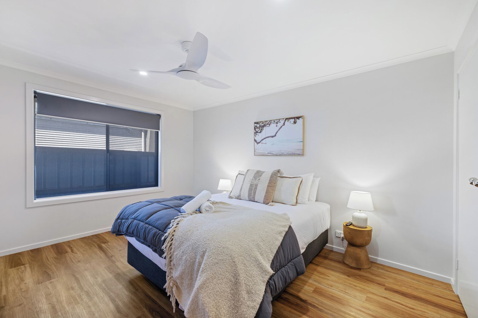 Une chambre à coucher comprenant un lit, un ventilateur de plafond, une fenêtre avec des stores, une table de chevet avec une lampe et un tableau accroché au mur.