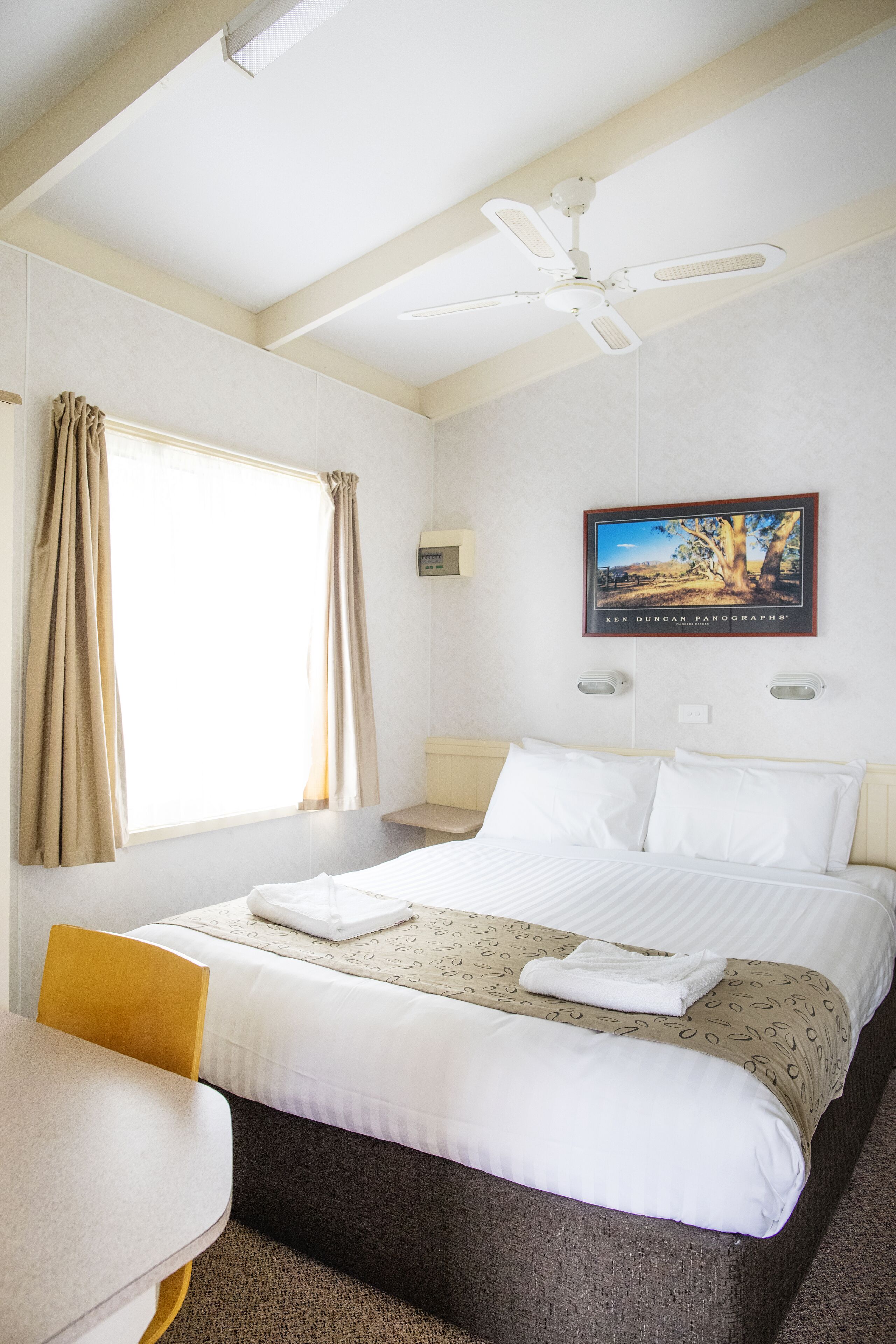 Habitación de hotel con cama, escritorio, silla, ventilador de techo, ventana con cortinas e imagen enmarcada en la pared.