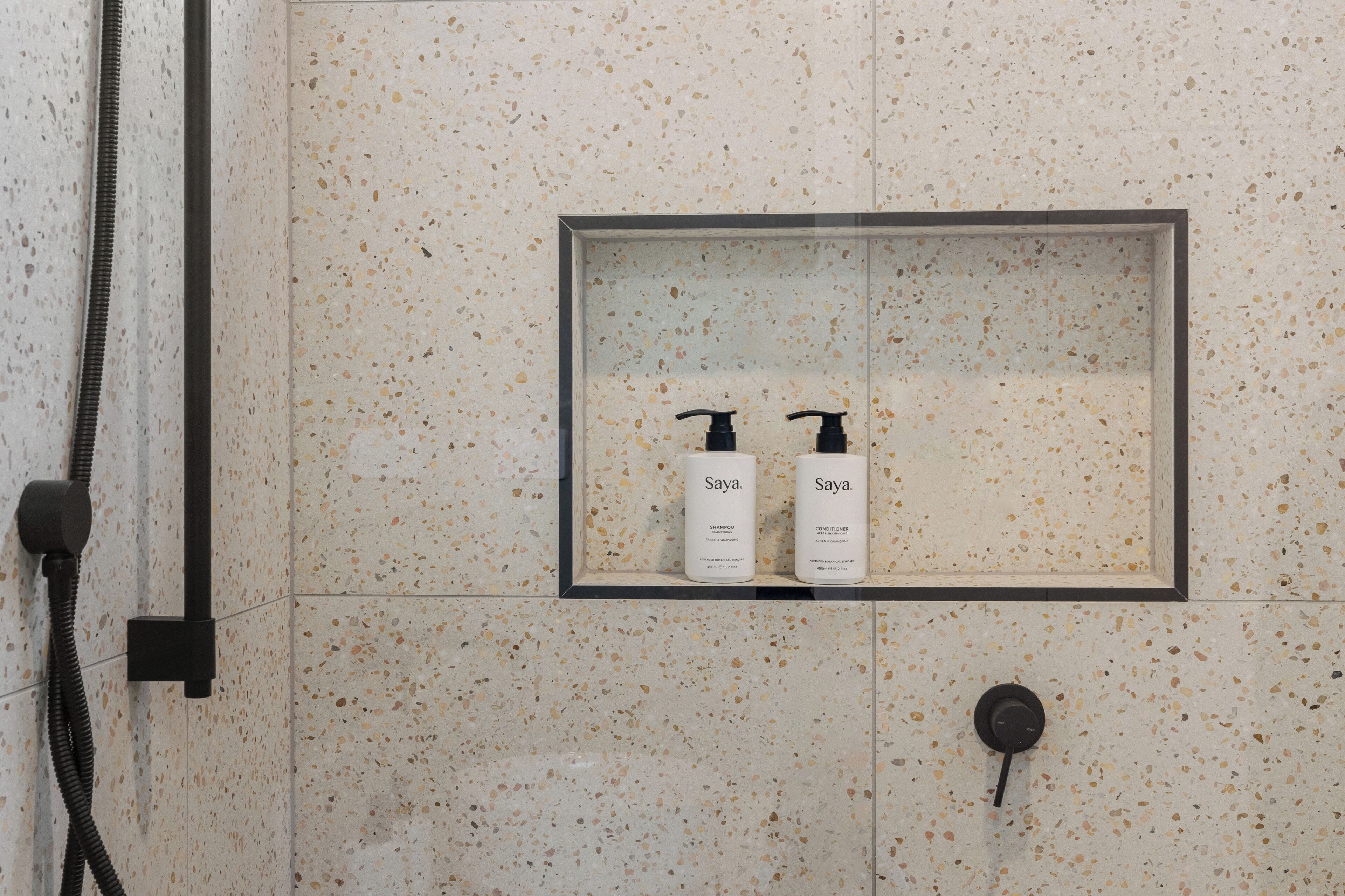 A bathroom with a speckled tile wall, a built-in shelf with two bottles of Soya shampoo and conditioner, and a showerhead.