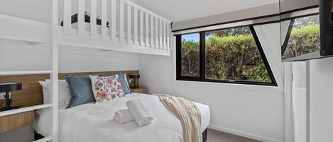A modern bedroom with a bunk bed, a large window with a view of greenery, a bedside table with a lamp, and a flat-screen TV mounted on the wall.