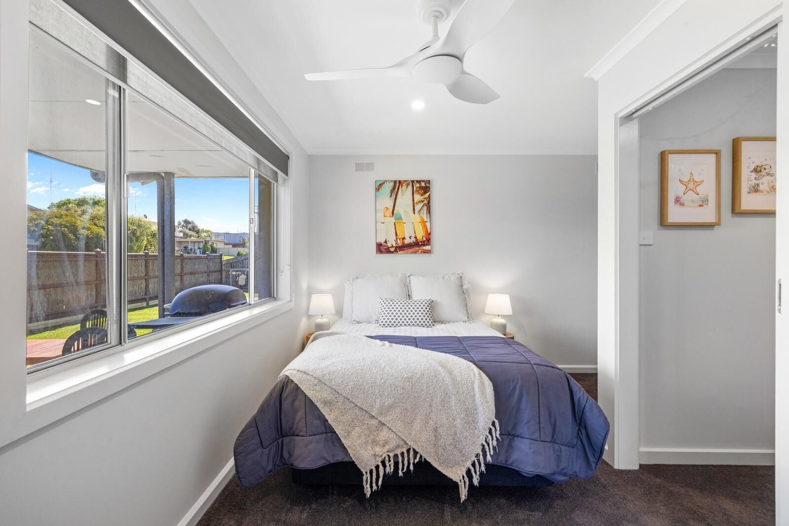 Un dormitorio con un ventanal grande, una cama con edredón azul y blanco, un ventilador de techo y un cuadro en la pared.
