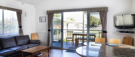 A modern living room with a sofa, coffee table, and dining area with chairs. There is a television mounted on the wall and a sliding glass door leading to an outdoor patio.