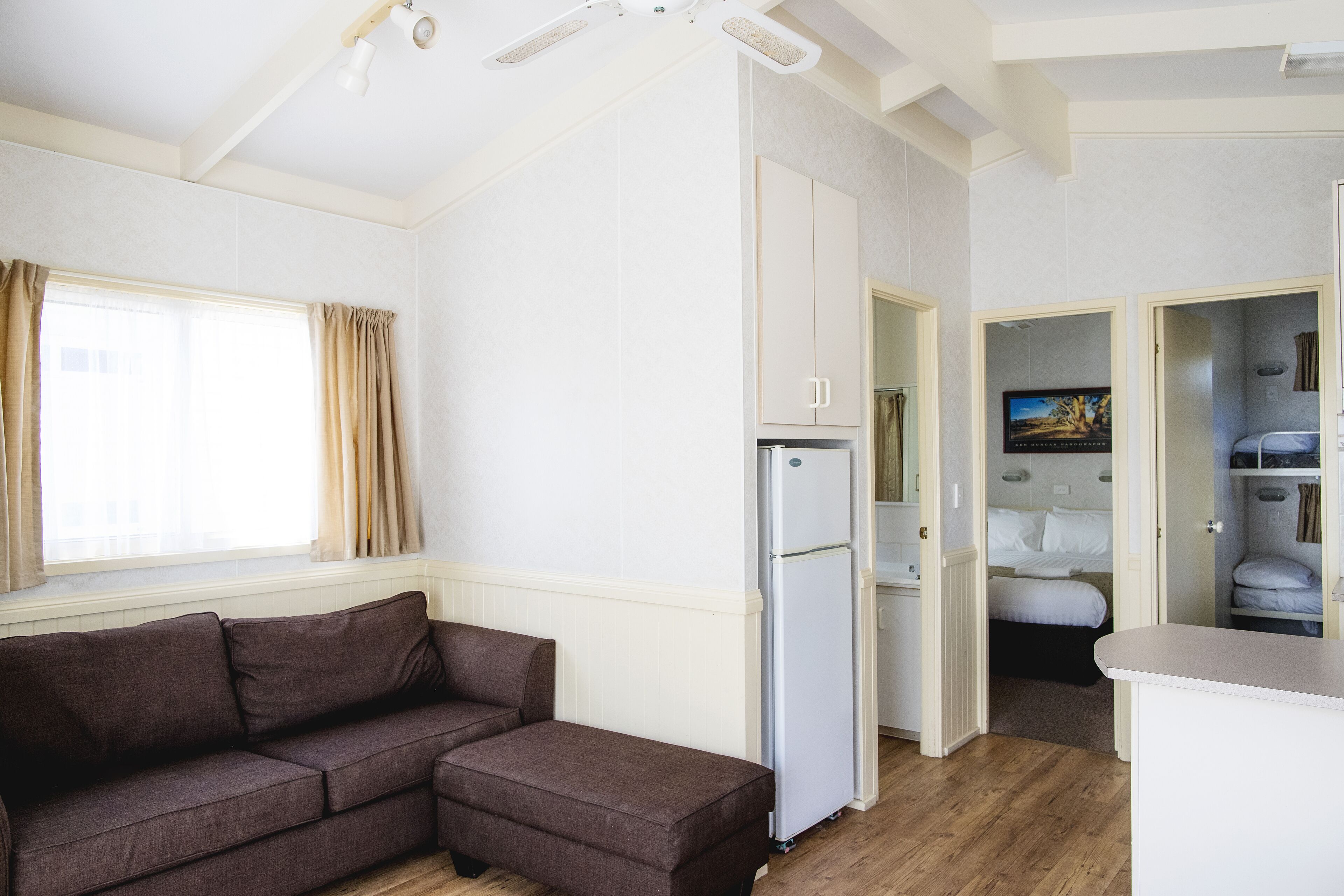 Quarto compacto com um sofá marrom, um refrigerador branco e uma cama ao fundo.