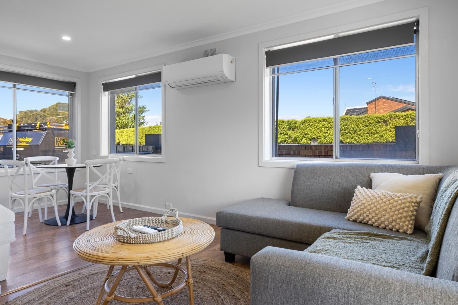 A modern living room with a grey sofa, white chairs, a round wicker table, and large windows offering a view of a green landscape and a building with solar panels.
