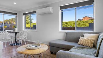 A modern living room with a grey sofa, white chairs, a round wicker table, and large windows offering a view of a green landscape and a building with solar panels.