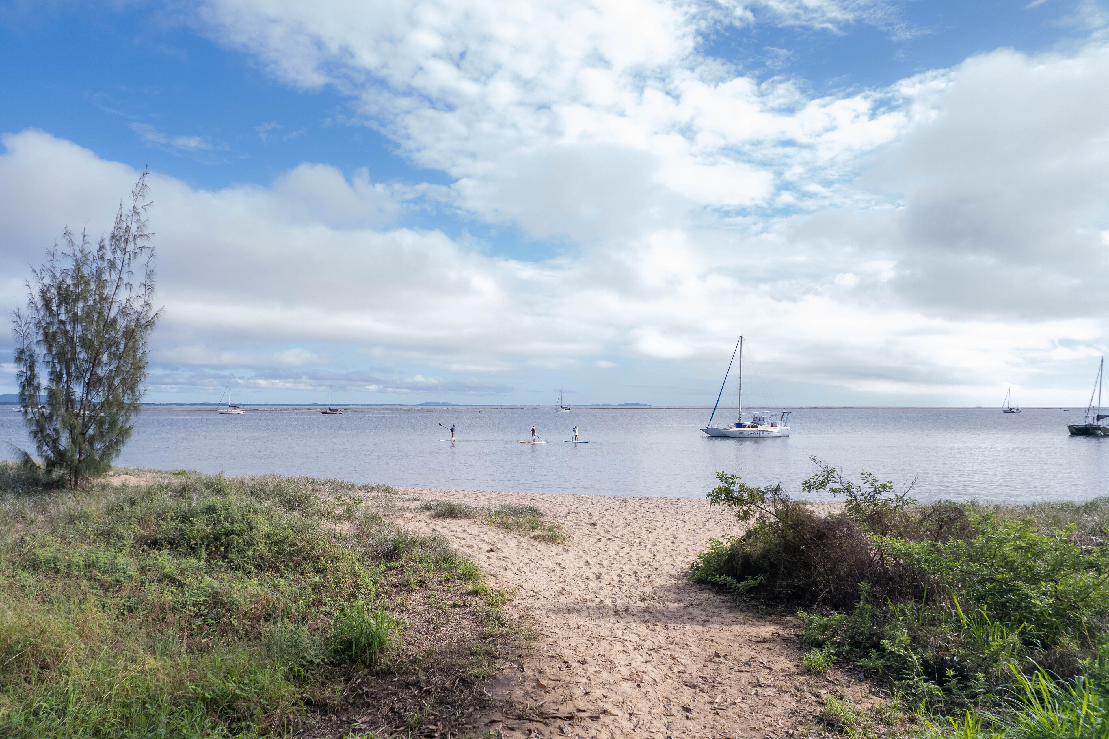 Una playa con un sendero arenoso que lleva al agua, barcos a lo lejos y un cielo despejado.