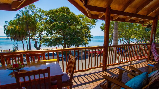 Een houten terras met tafel en stoelen, met uitzicht op een strand met palmbomen en de oceaan.