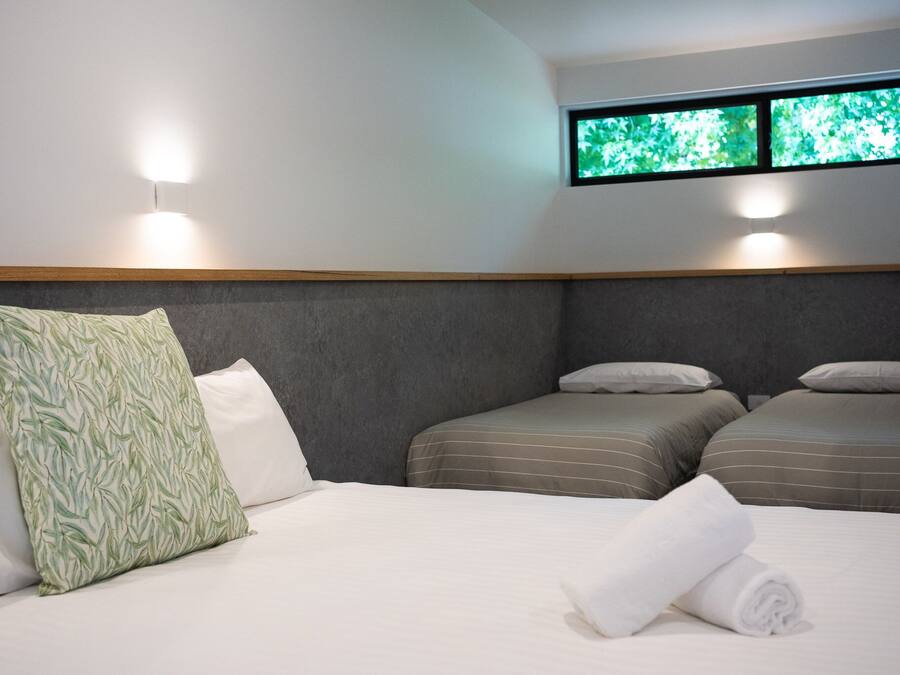 Habitació d'hotel amb dos llits individuals, un llit gran amb una tovallola arrotllada, i una finestra amb vista a la vegetació.