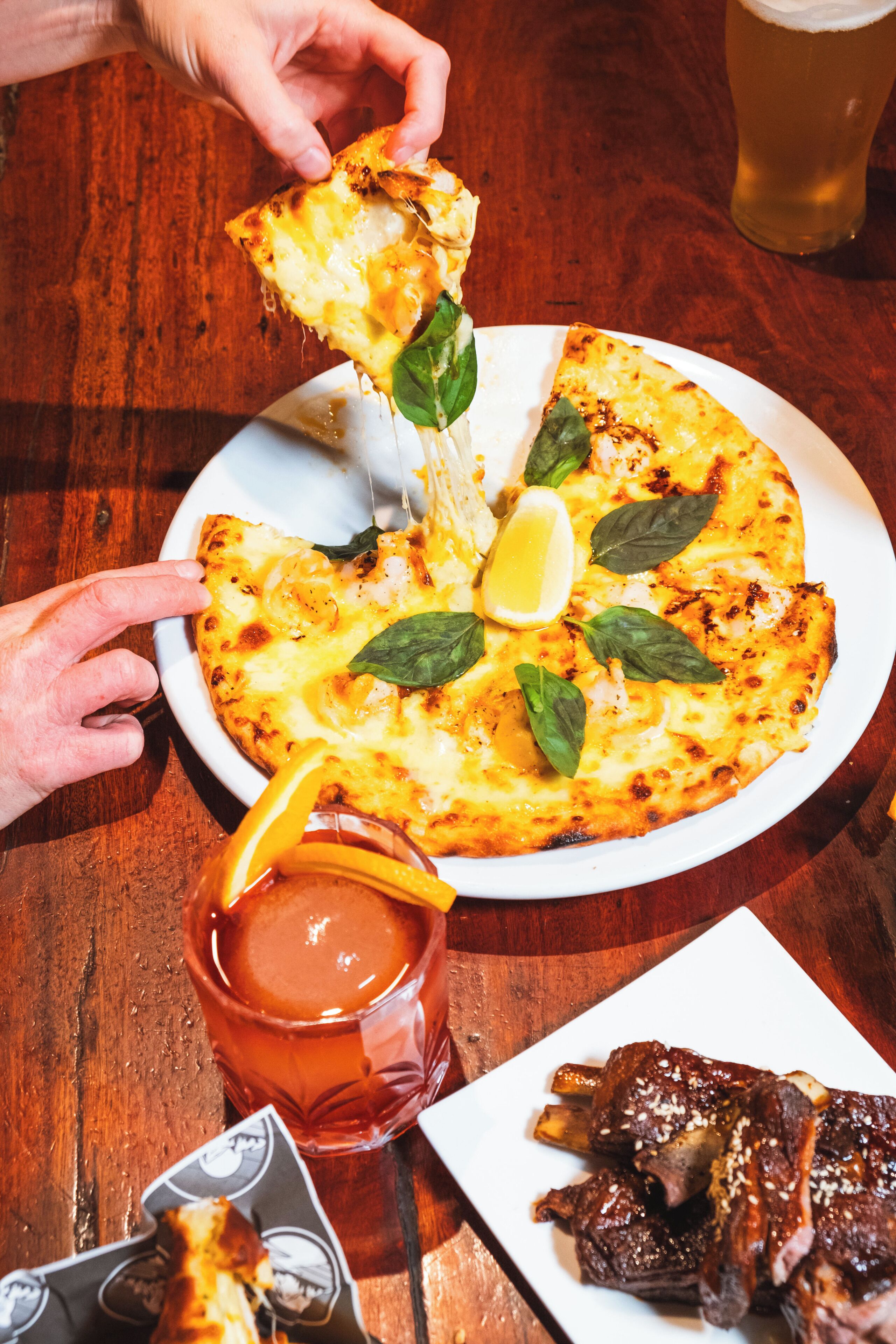 Pizza met kaas en basilicumblaadjes op een wit bord, een glas rode drank, en een bord met spareribs.