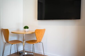 Ein kleiner, runder Holztisch mit zwei Stühlen, einem Blumentopf und einem an der Wand befestigten Flachbildfernseher.