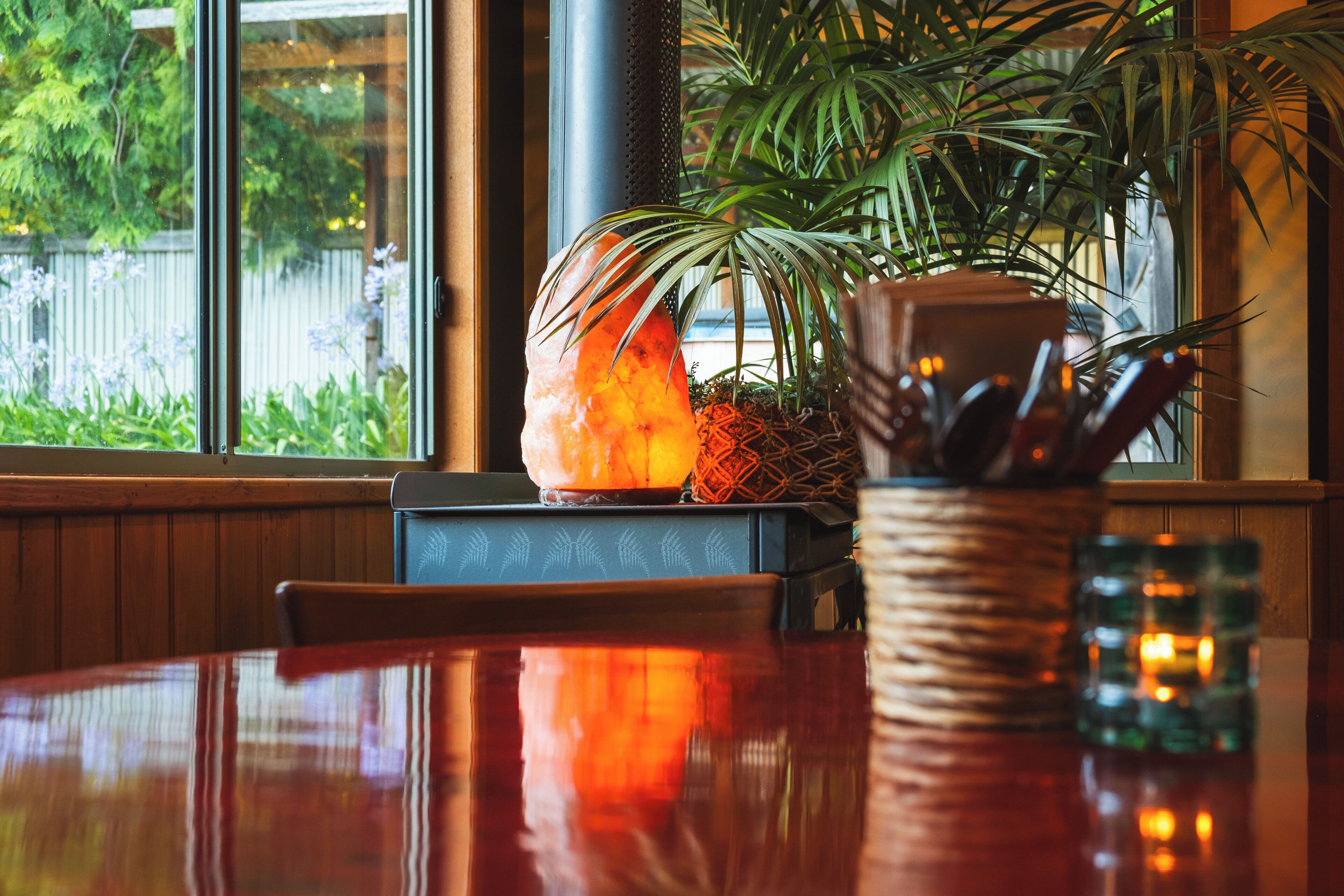 A restaurant interior with a wooden table, chairs, a salt lamp, a potted plant, and a window with a view of greenery.