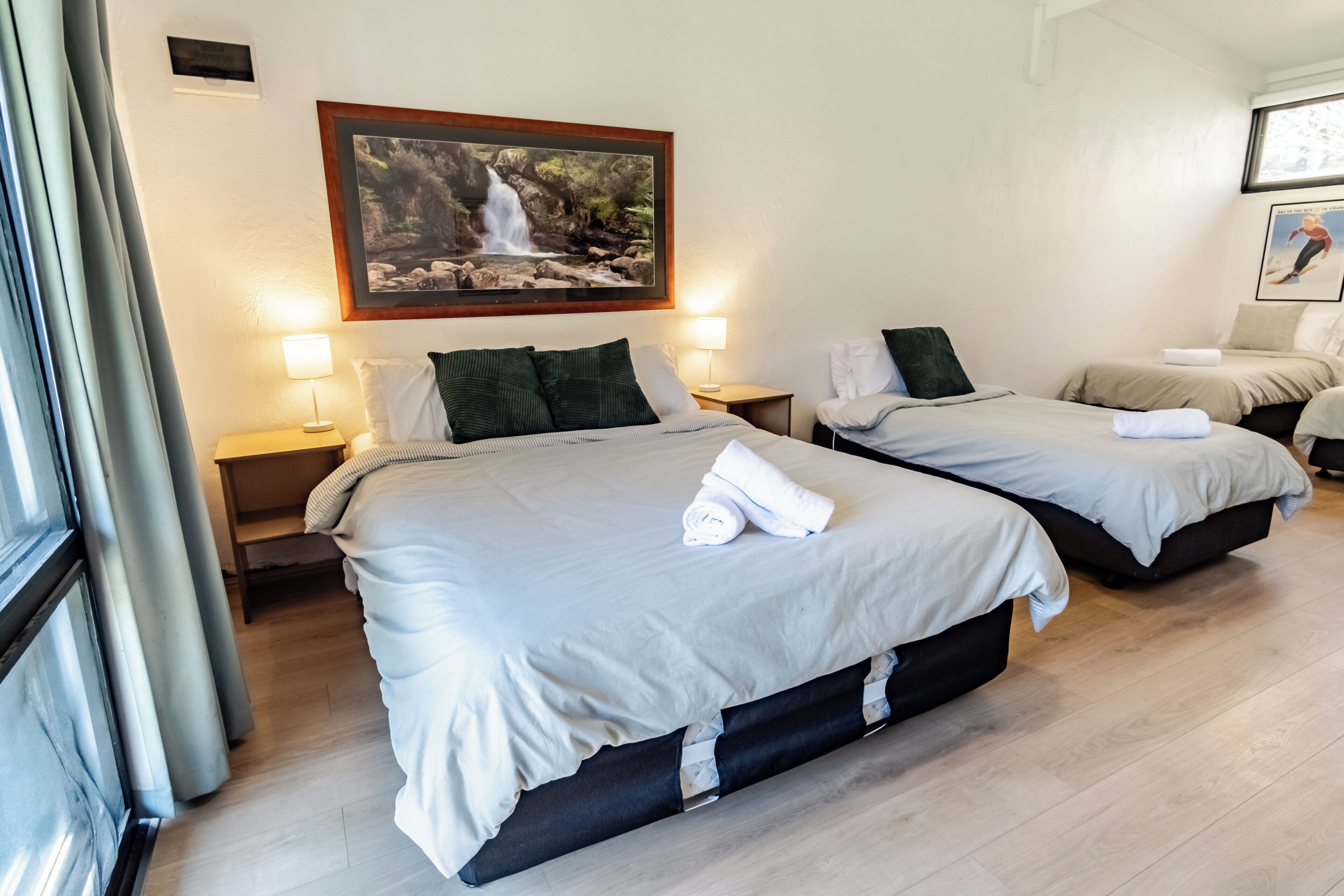 A hotel room with two beds, a painting of a waterfall, and a window with curtains.