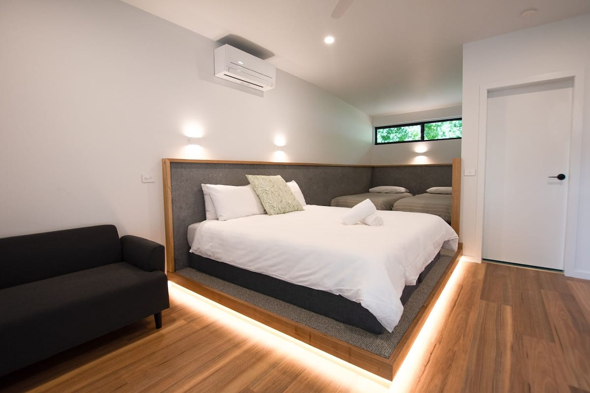 A modern bedroom with a large bed, a dark sofa, and a window with a view of greenery.
