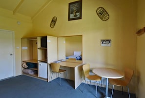 Une petite pièce comprenant un lit, un bureau et deux chaises. Les murs sont décorés et il y a une porte.