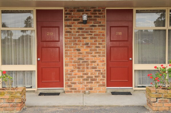 A two-story building with red doors numbered 212 and 211, a brick wall, and a small brick planter with flowers. - The Peninsula Motor Inn (Tyabb)