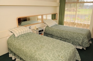 Quarto com duas camas de solteiro, uma pequena mesa de cabeceira com abajur e janela com vista para o verde.
