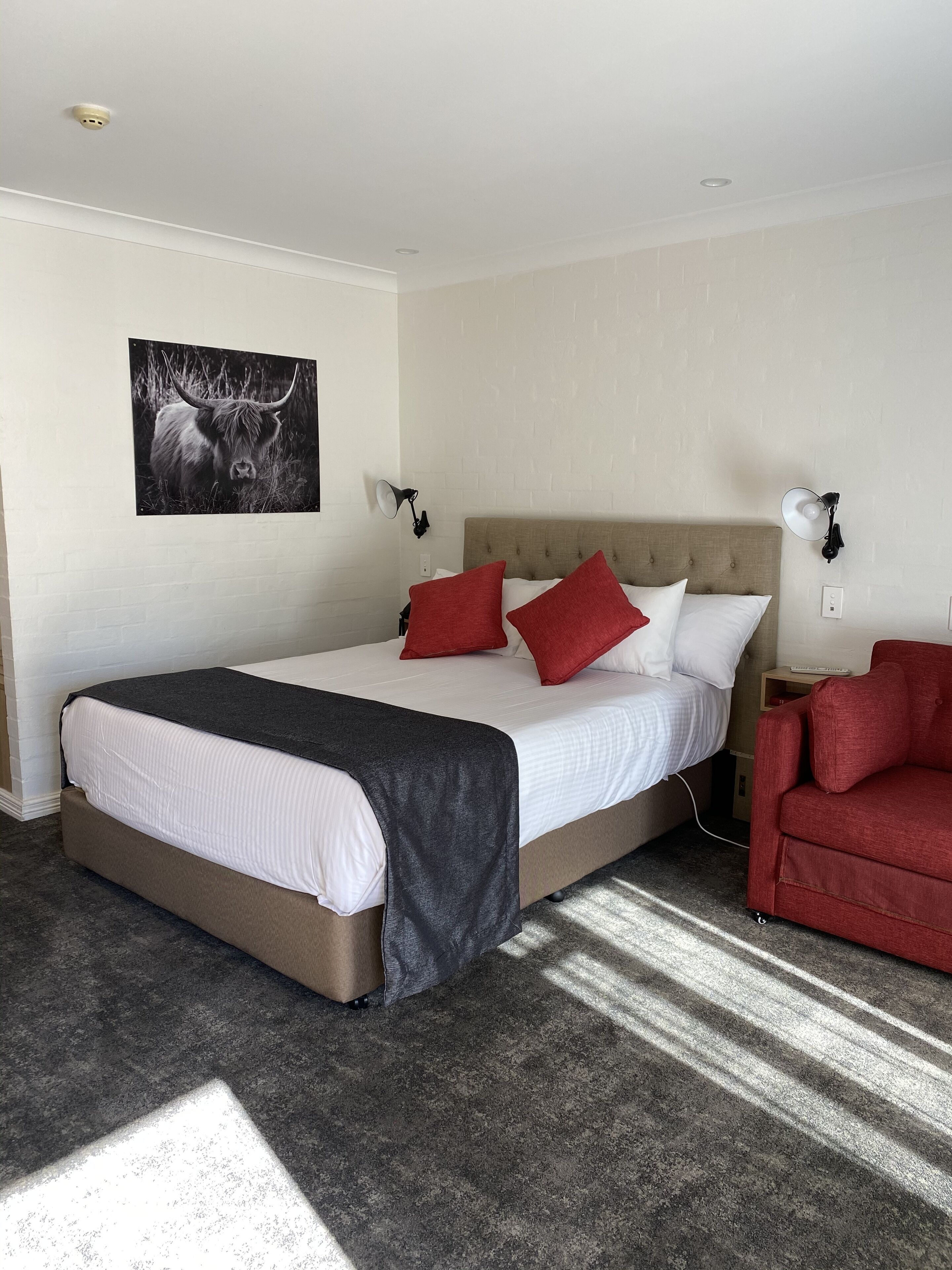 A neatly made bed with a grey blanket, red pillows, and a headboard. There is a red armchair and a wall-mounted lamp beside the bed.