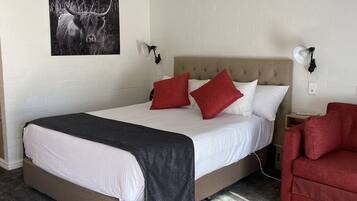 A neatly made bed with a grey blanket, red pillows, and a headboard. There is a red armchair and a wall-mounted lamp beside the bed.