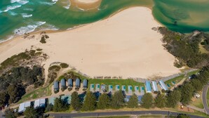 Vista aérea de una playa con aguas cristalinas, una orilla arenosa y una hilera de casas móviles con techo azul.