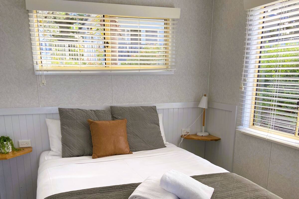A neatly made bed with a grey quilt, white linens, and a folded towel. There are two bedside tables with lamps, a window with blinds, and a small shelf with a plant.