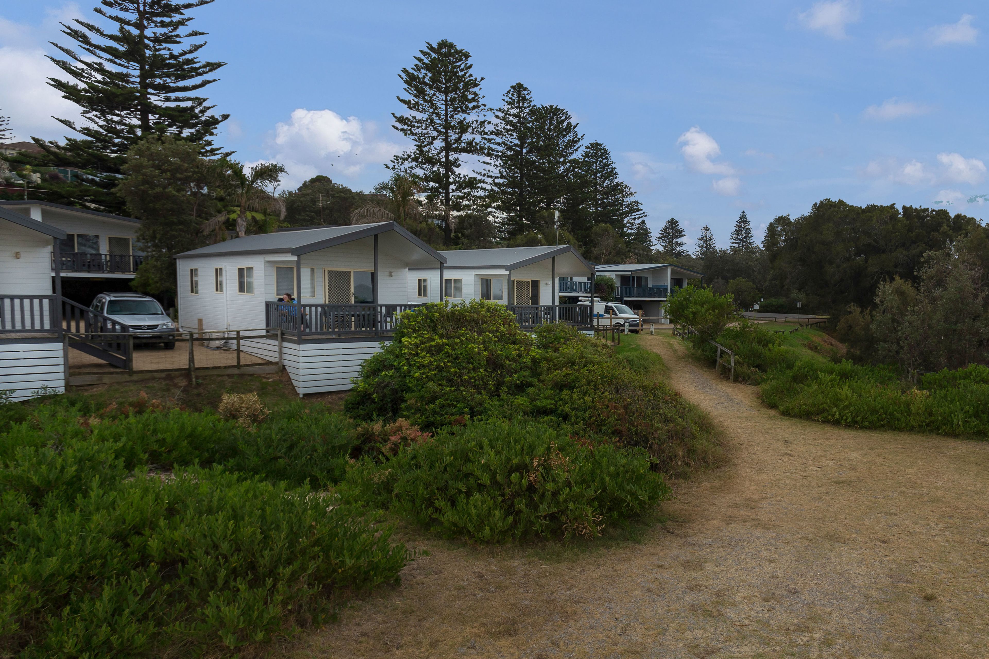 A row of white mobile homes with balconies, surrounded by greenery and a dirt path.