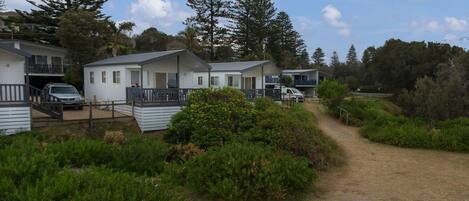 A row of white mobile homes with balconies, surrounded by greenery and a dirt path.