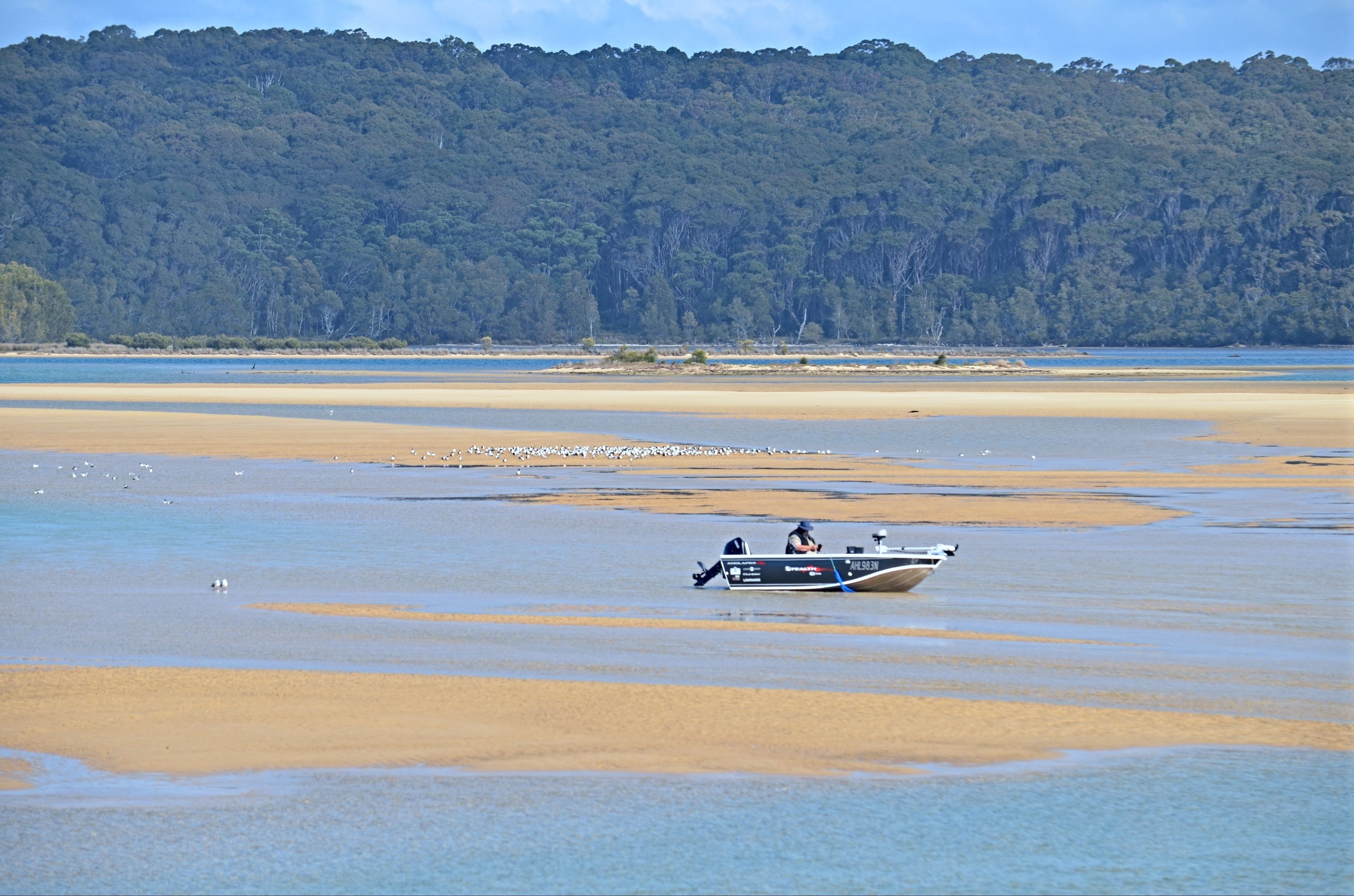 A boat on a wide, shallow water body with a forested hill in the background.