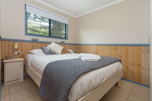 Een slaapkamer met een bed, nachtkastje, raam en houten lambrisering.