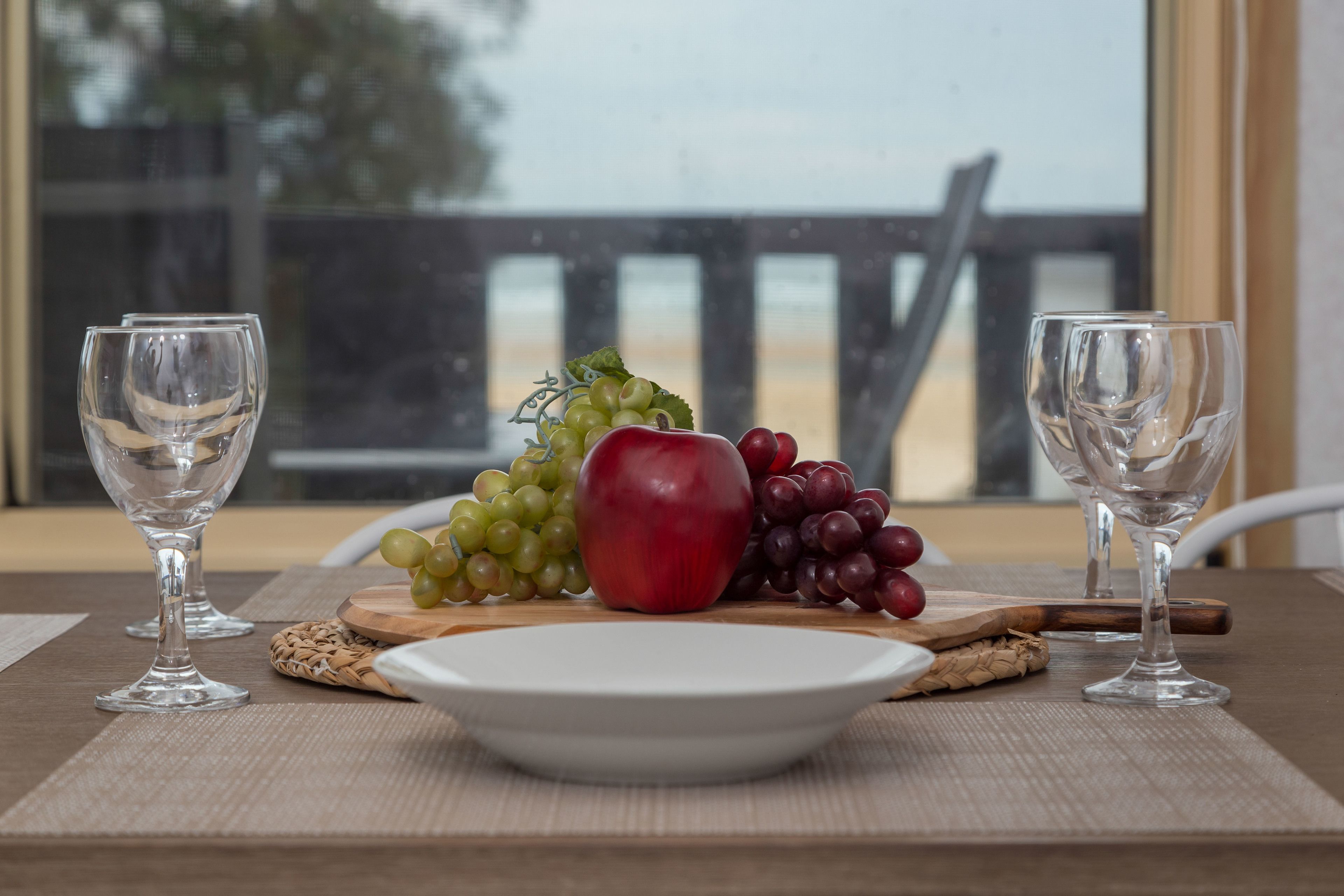 Una mesa con un plato de fruta, dos copas de vino vacías y una tabla de madera.