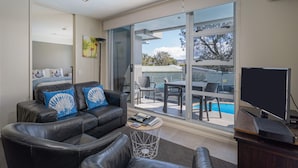 Una sala moderna con un sofá de cuero negro, una mesa de centro y un televisor. Hay una puerta corrediza de vidrio que da a un área exterior con piscina y mobiliario de patio.