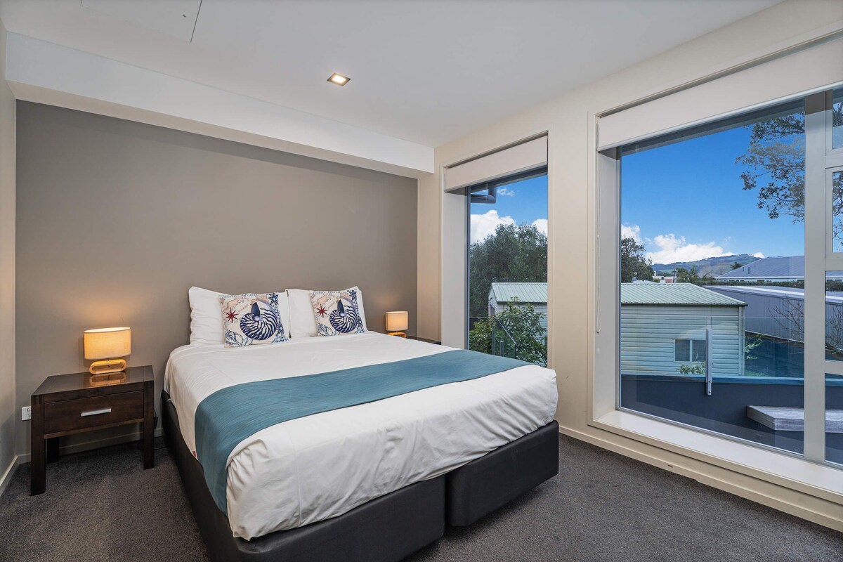 A bedroom with a large window, a bed with a white and teal bedspread, a nightstand with a lamp, and a view of houses outside.