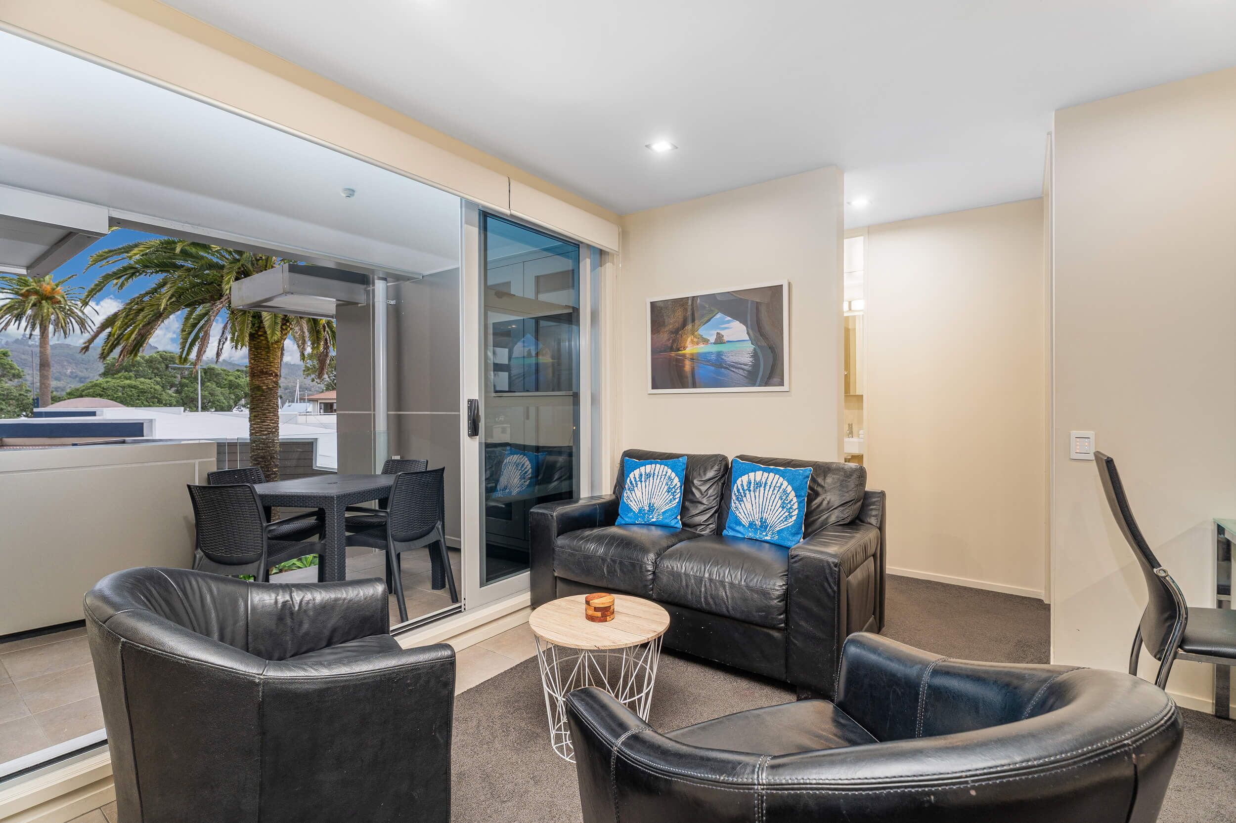 A modern living room with a glass door leading to a balcony, a black leather sofa with blue and white cushions, and a small round coffee table.