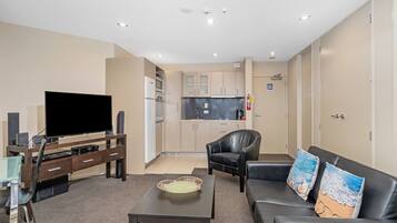 Una sala moderna con una televisión de pantalla plana, una mesa de centro, un sofá con cojines con motivos playeros, un sillón de cuero y una zona de cocina al fondo.