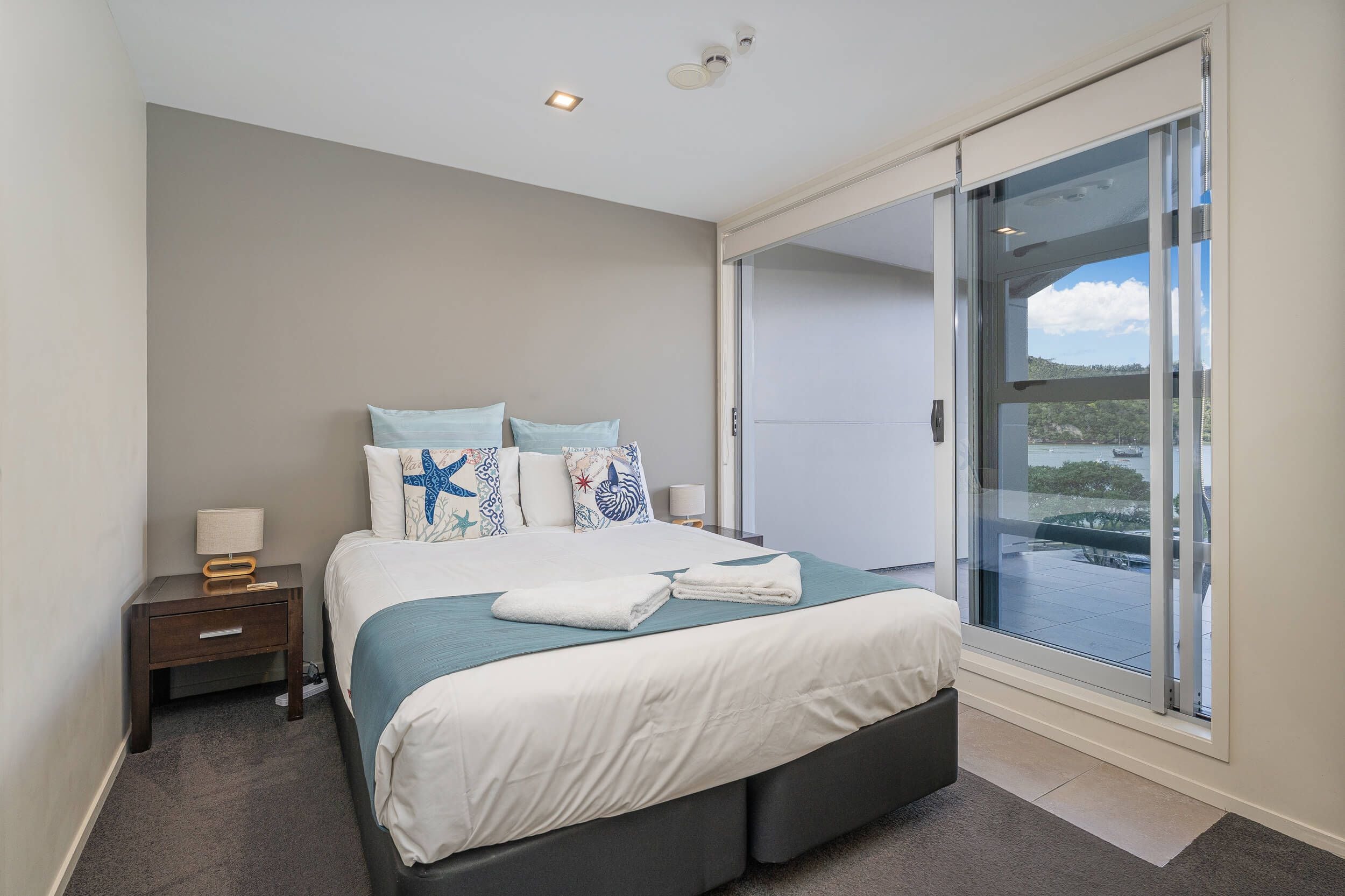 A modern bedroom with a large bed, a nightstand, a lamp, and a sliding glass door leading to a balcony.