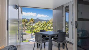 A balcony with a table and chairs, a sliding glass door, and a view of a green landscape with palm trees and mountains.