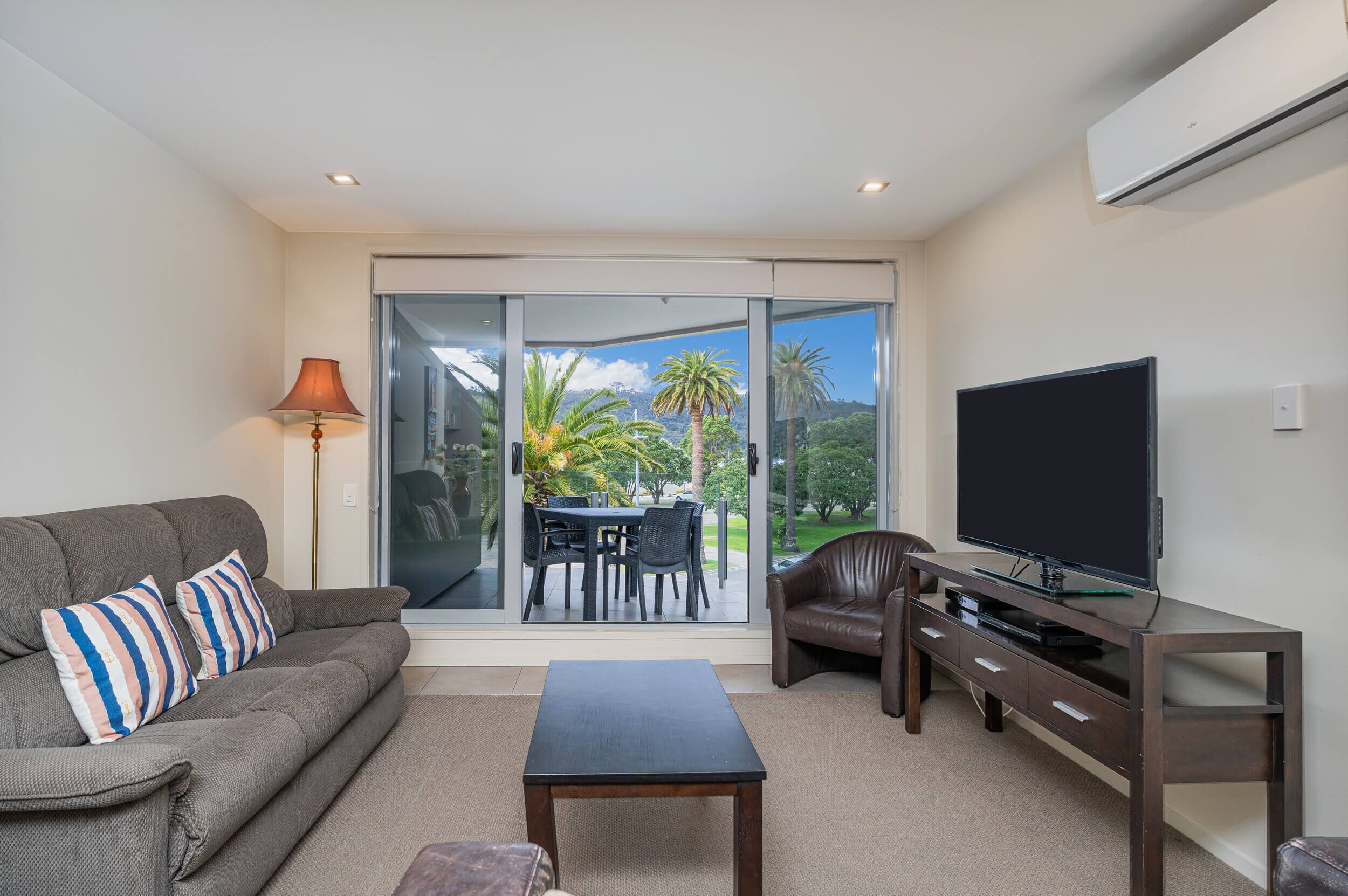 A living room with a sofa, a coffee table, a TV on a wooden stand, and a sliding glass door leading to an outdoor area with a table and chairs.