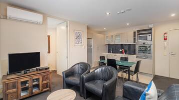 A modern living room with a flat-screen TV, leather armchairs, a small round table, and a sofa. A kitchen with a dining area is visible in the background.