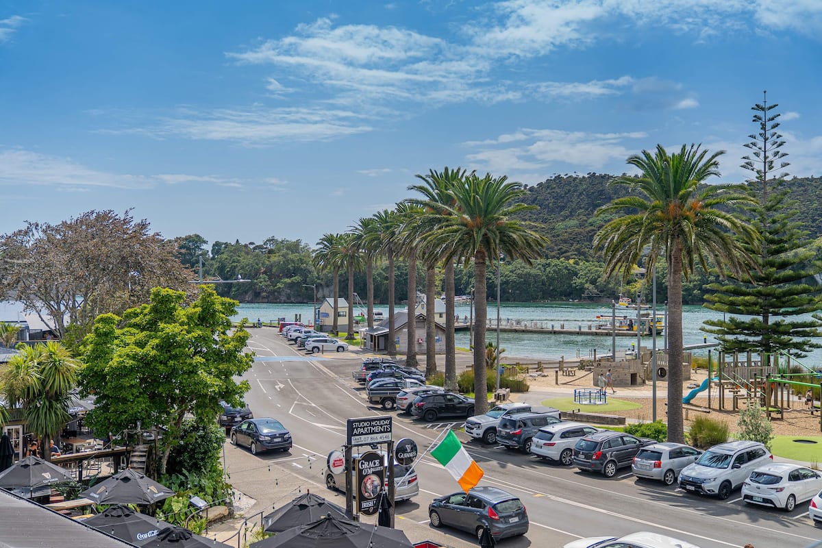 Een parkeerplaats met auto's, een bord met 'Studio Holiday Apartments' en uitzicht op een strand met palmbomen en een speeltuin.