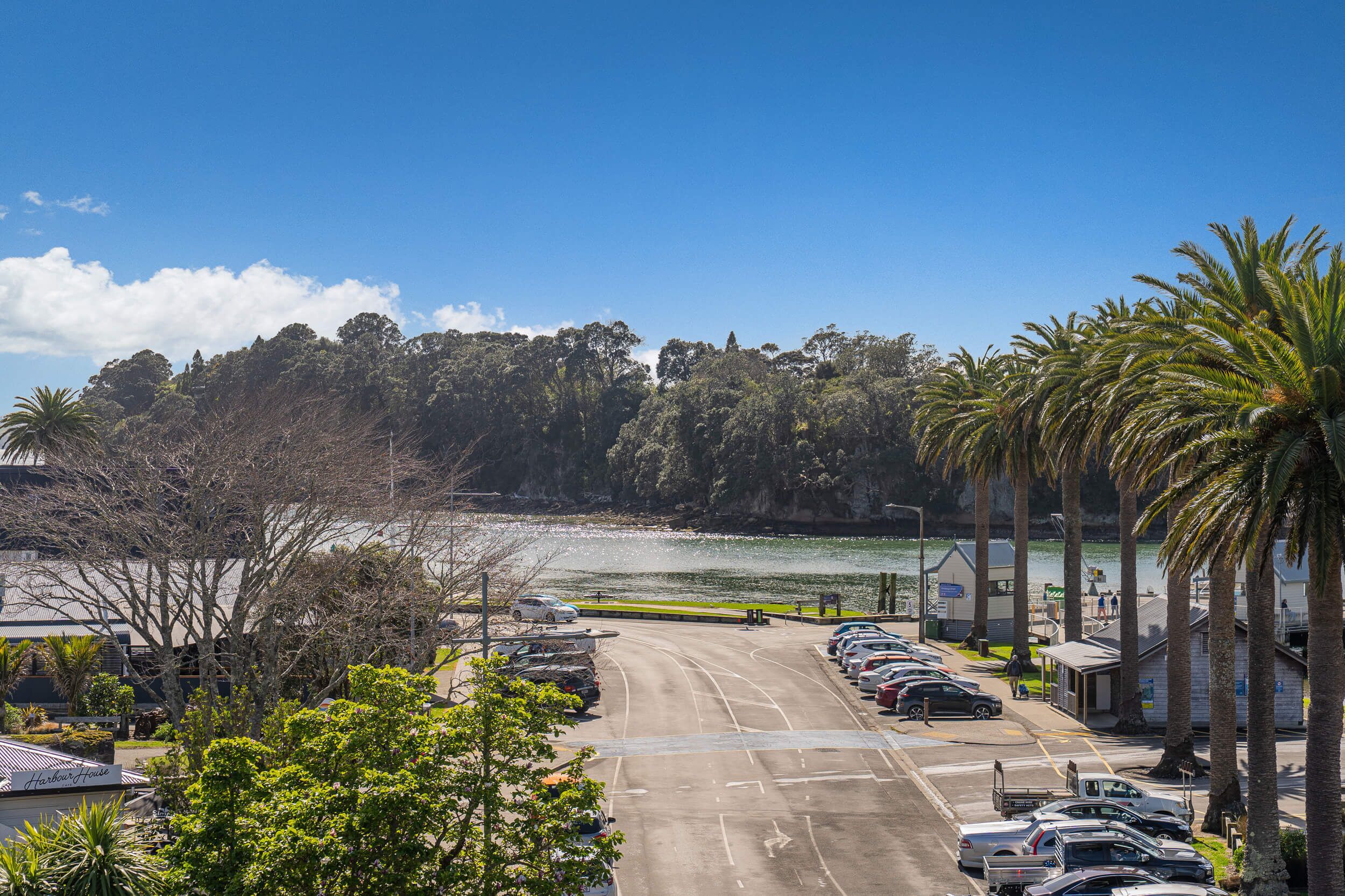 Un parking rempli de voitures, un bâtiment portant l’inscription « Harbor House », et une vue sur une étendue d’eau et des arbres.