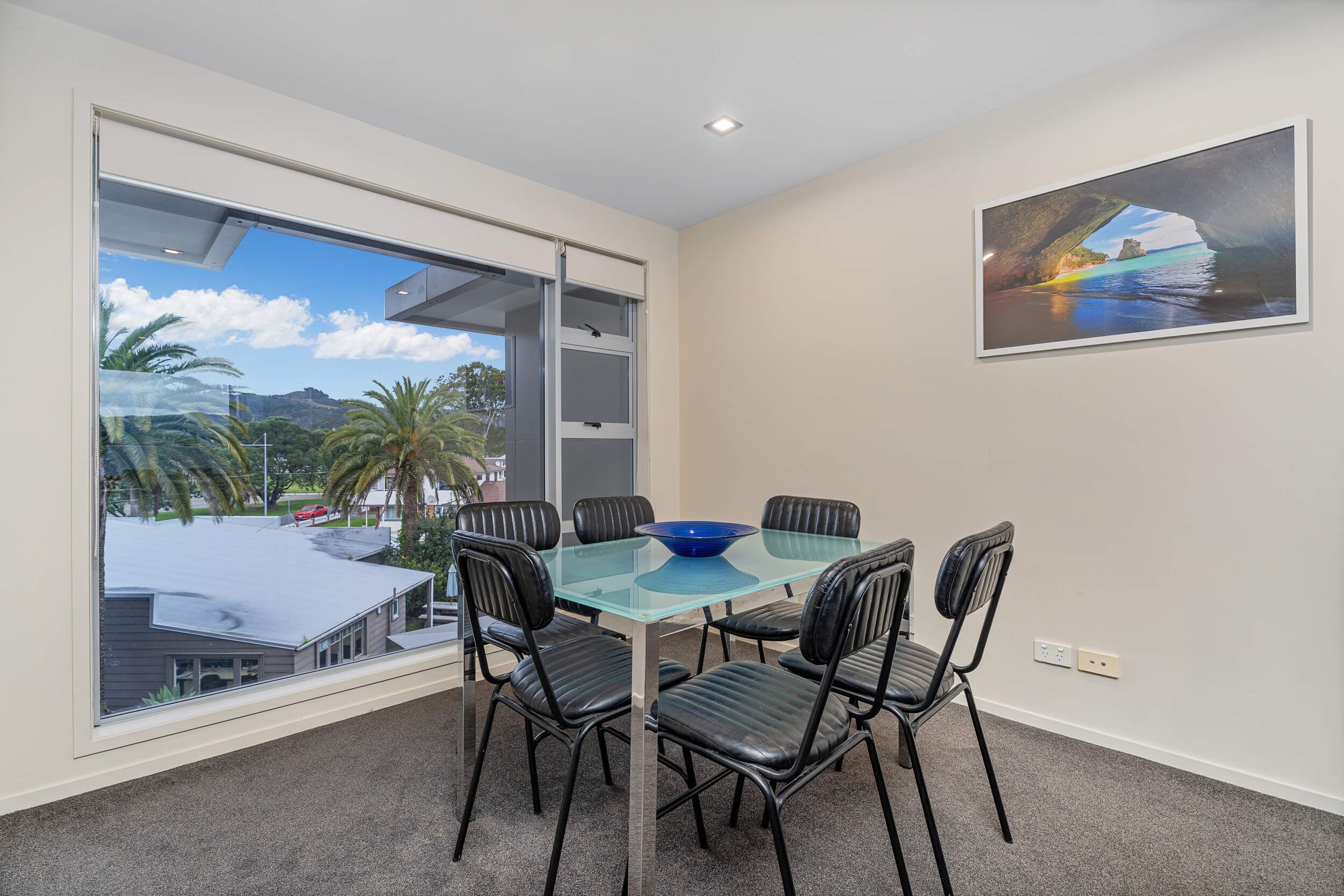 A room with a glass table and chairs, a painting on the wall, and a view of palm trees and a building outside.