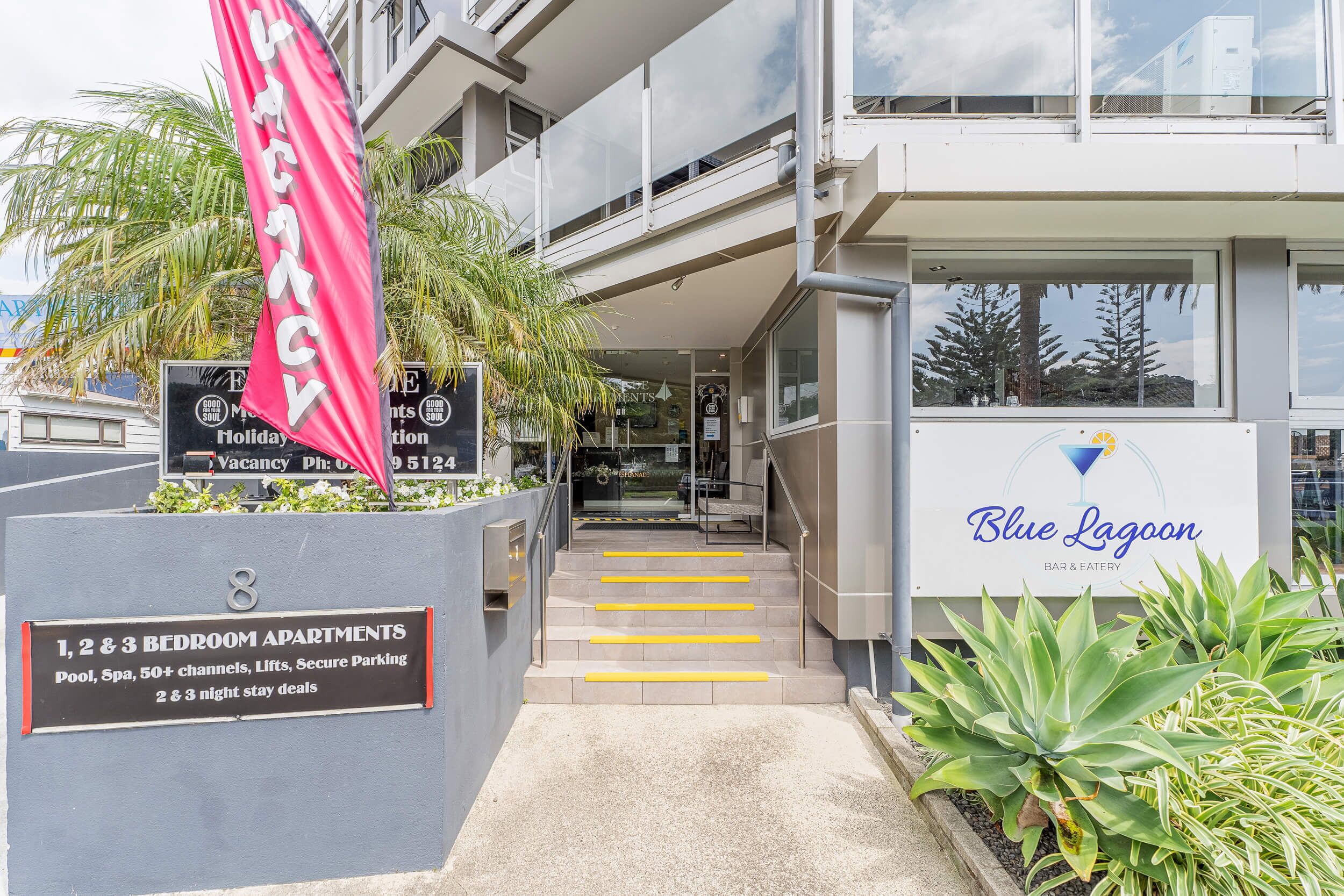 A building with a sign for 'Blue Lagoon' bar and eatery, and a sign for 'Vacancy' with a phone number. There are 1, 2, and 3 bedroom apartments available, with amenities like pool, spa, 50+ channels, lifts, and secure parking.